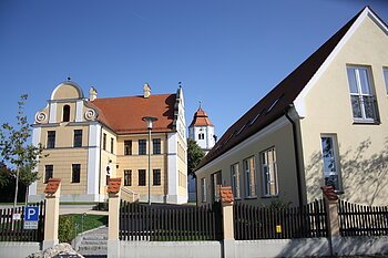 Gelbes historisches Gebäude und modernes Haus mit roten Dächern unter blauem Himmel, im Vordergrund Zaun und Parkplatzschild.