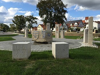 Steinbrunnen mit Wappen und Steinskulpturen auf gepflastertem Platz in Wohngebiet bei bewölktem Himmel