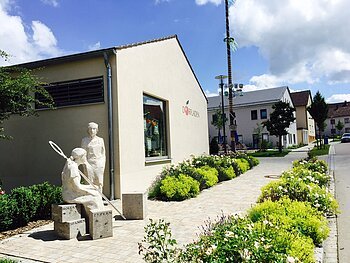 Skulptur von zwei Figuren vor einem Gebäude mit Schriftzug „Dorfladen“ und bepflanztem Weg bei sonnigem Himmel.