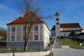 Gelbes Wohnhaus mit Baum davor und Kirche mit Treppe bei blauem Himmel im Hintergrund.