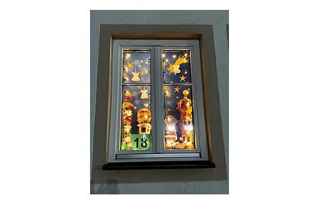 Adventskalenderfenster 18.12.2024 - Schule & OGTS Adventskalenderfenster mit einer Umrandung in beige weihnachtlich dekoriert. Darin mit Weihnachtsbeleuchtung beleuchtete Engelsfiguren, eine Sternenschnuppe und Sternen. Unten links die Zahl 18 auf grünem Hintergrund.