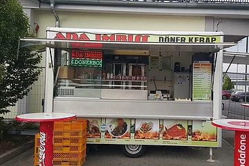 Imbisswagen mit geöffnetem Verkaufsfenster und zwei Stehtischen davor. Auf dem Wagen oben die Beschriftung „ADA Imbiss - Döner Kebab“ sichtbar unter dem geöffnetem Fenster verschiedene Abbildungen der erhältlichen Gerichte abgebildet. Links im Bild ein Baum, rechts ein Teil eines Parkplatzes zu sehen.
