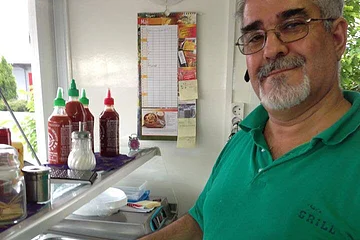 Mann in grünem Poloshirt und mit Brille hält einen auf Alufolie ausgebreiteten Dürüm belegt mit Salat, Tomaten und Fleisch in einer Imbissküche. Hinter ihm sind Edelstahlbehälter mit Beilagen zu sehen und verschiedene Zutaten wie Soßen und Gewürze. Im Hintergrund an der Wand hängt eine Uhr und ein Kalender.