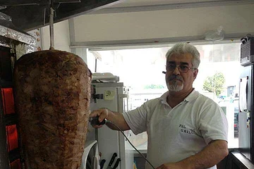 Ein Mann mit Brille, im weißen Hemd und einer schwarzen Schürze schneidet Fleisch von einem großen Döner-Spieß in einer Imbissküche, umgeben von weiteren Küchengeräten. Unter dem Spieß liegt bereits abgeschnittenes Fleisch.