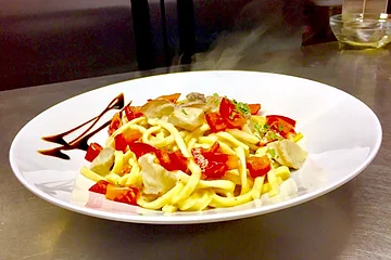 Teller mit dampfender Pasta, Tomatenstücken, Pilzen und Kräutern, dekoriert mit Balsamico-Soße steht auf einem braunem Holztisch.