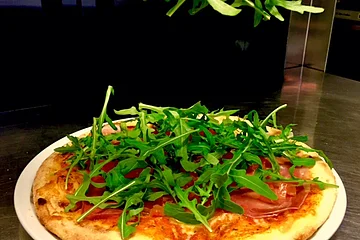 Hand legt Rucola auf eine belegte Pizza mit Schinken auf weißem Teller in Küche.