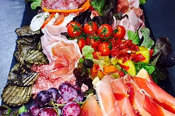 Antipasti-Platte auf schwarzer Unterlage mit Trauben, Tomaten, Melone, verschiedenen Wurst- und Käsesorten sowie gegrilltem Gemüse auf Salatblättern.