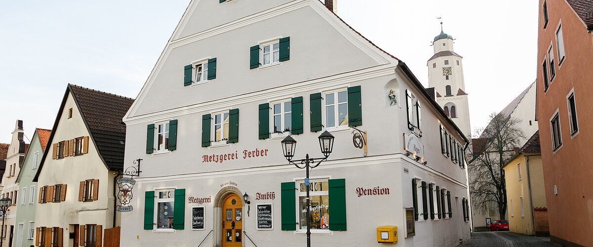 Graues mehrstöckiges Gebäude mit grünen Fensterläden, einer Treppe mit Geländern und dem Schriftzug "Metzgerei Ferber" oberhalb der Eingangstür, an einer gepflasterten Straße. Links von der Tür der Schriftzug "Metzgerei", rechts davon "Imbiss" und "Pension" an der Fassade. Ein gelber Briefkasten hängt an der vorderen Fassade.