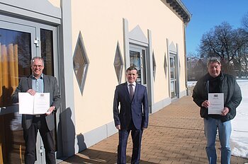 Drei Männer stehen vor einem hellen Gebäude auf einem gepflastertem Platz, zwei halten Urkunden in den Händen, Schnee rechts im Bild liegt auf dem Boden.