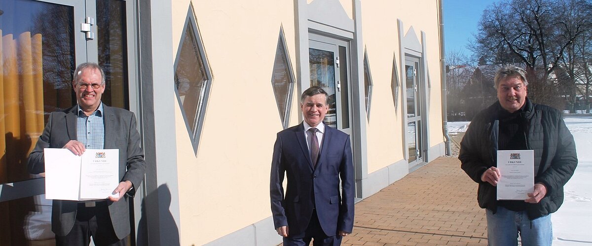 Drei Männer stehen vor einem hellen Gebäude auf einem gepflastertem Platz, zwei halten Urkunden in den Händen, Schnee rechts im Bild liegt auf dem Boden.