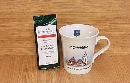 Geschenkidee der Stadt Monheim: eine weiße Tasse mit Monheim-Motiv und der Aufschrift "Monheim - Die Drei-Stämme-Stadt" rechts im Bild. Links davon eine Teeverpackung mit der Mischung und dem Text „Monheimer Früchtekorb“ stehen vor einer Holzoberfläche.