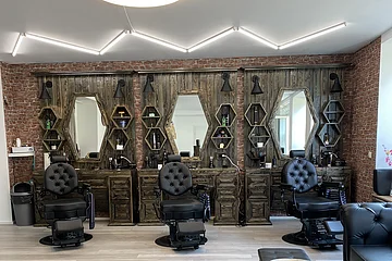 Friseursalon mit drei schwarzen Friseurstühlen vor dunkelbraunen Holzspiegeln und Backsteinwand. Rechts im Bild kleiner Ausschnitt vom Sitzbereich mit schwarzen Möbeln. An der Decke moderne Leuchte.