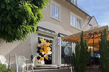 Außentreppe zu einem Eingang, daneben ein Eis-Café. An der Tür hängen gelb-schwarz-weiße Luftballons. Rechts davon der Sitzbereich des Eis-Cafés mit einem orangen Sonnenschirm. Hinter dem Gebäude ein strahlend blauer Himmel.
