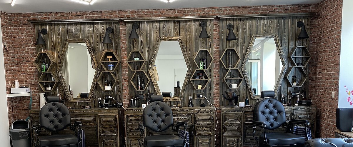 Friseursalon mit drei schwarzen Friseurstühlen vor dunkelbraunen Holzspiegeln und Backsteinwand. Rechts im Bild kleiner Ausschnitt vom Sitzbereich mit schwarzen Möbeln. An der Decke moderne Leuchte.