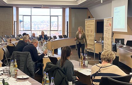 Seminarraum mit hellen Tischen und schwarzen Stühlen, mehreren Personen sitzen an den Tischen, eine Frau präsentiert vor Flipcharts und einem Projektorbild.