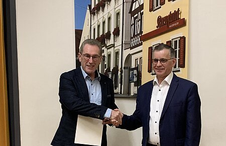 Zwei Männer in Anzügen schütteln sich die Hand vor einer Wand mit einem Bild von Fachwerkhäusern. Der Mann links hält eine Urkunde in der anderen Hand.