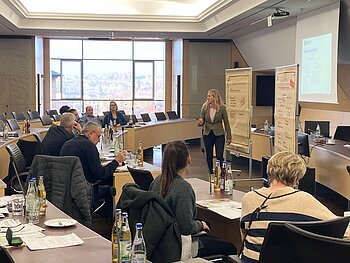 Seminarraum mit hellen Tischen und schwarzen Stühlen, mehreren Personen sitzen an den Tischen, eine Frau präsentiert vor Flipcharts und einem Projektorbild.