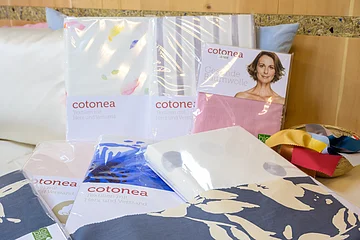 Mehrere verpackte Textilien der Marke "Cotonea" auf einem weißen Sofa, daneben eine Schale mit bunten Stoffmustern, im Hintergrund eine Holzwand.