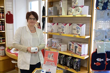 Apothekerin in weißem Kittel hält medizinisches Blutdruckmessgerät vor einem Regal mit weitern Blutdruckmessgeräten in Apotheke. Rechts im Bild Wärmeflaschen und Wärmekissen.