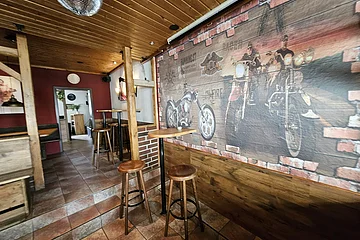 Innenraum einer Bar mit Holzdecke, hohen Stehtischen und Barhockern. Rechts an der Wand ein großes Wandbild mit Motorrädern und Ziegelsteinmuster. Hinten ein weiterer Raum zu sehen.
