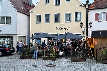 Außenbereich einer Bar mit Gästen an Tischen sitzend und unter zwei großen schwarzen Sonnenschirmen stehend, vor einem gelben Gebäude mit Schriftzug „Nest“.
