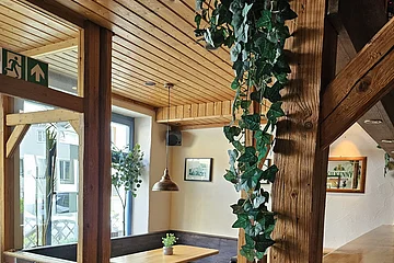 Im Vordergrund eine Holztheke mit hohen Barstühlen, einer hängender Pflanze. Im Hintergrund ein Sitzbereich mit Tisch und schwarzer Sitzbank, Fenster, Bildern an der Wand und Notausgangsschild zu sehen.