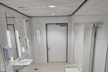 Barrierefreies Badezimmer mit Toilette, Waschbecken, Haltegriffen, Spiegel, Heizung und Sitzgelegenheit, alles in weiß/weiß gefließt.