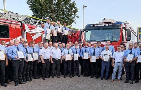 Gruppenfoto von Feuerwehrleuten in blauer Uniform in mehreren Reihen vor zwei roten Feuerwehrfahrzeugen, die meisten halten Urkunden, bei blauem Himmel.