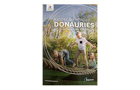 Titelseite der Broschüre "Kinder & Familien Donauries" - Ein himmlischer Glückstreffer vom Ferienland Donau-Ries. Auf dem Titelbild drei Kinder spielend auf einem Kletterseil im Freien, umgeben von Bäumen und Wiese.