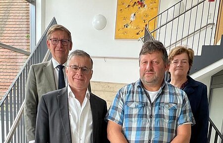 Vier Personen stehen blickend in die Kamera auf einer Treppe in einem Gebäude mit schwarzem Geländer und großem Fenster. An der Wand hängt ein gelbes Gemälde.