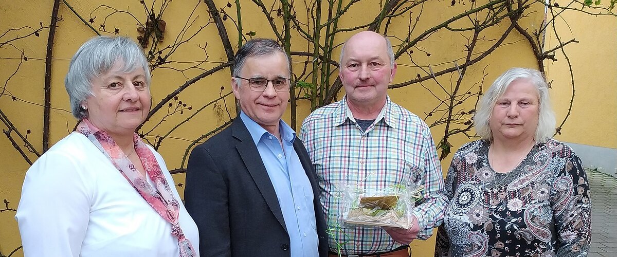 Vier Personen stehen nebeneinander vor einer gelben Wand mit kahlen Ästen, eine Person hält ein verpacktes Geschenk.