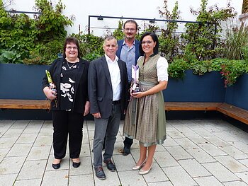 Vier Personen stehen auf gepflastertem Hof vor einem schwarzen Hochbeet mit grünen Pflanzen und Bänken entlang des Beetes, zwei Personen halten Geschenke.