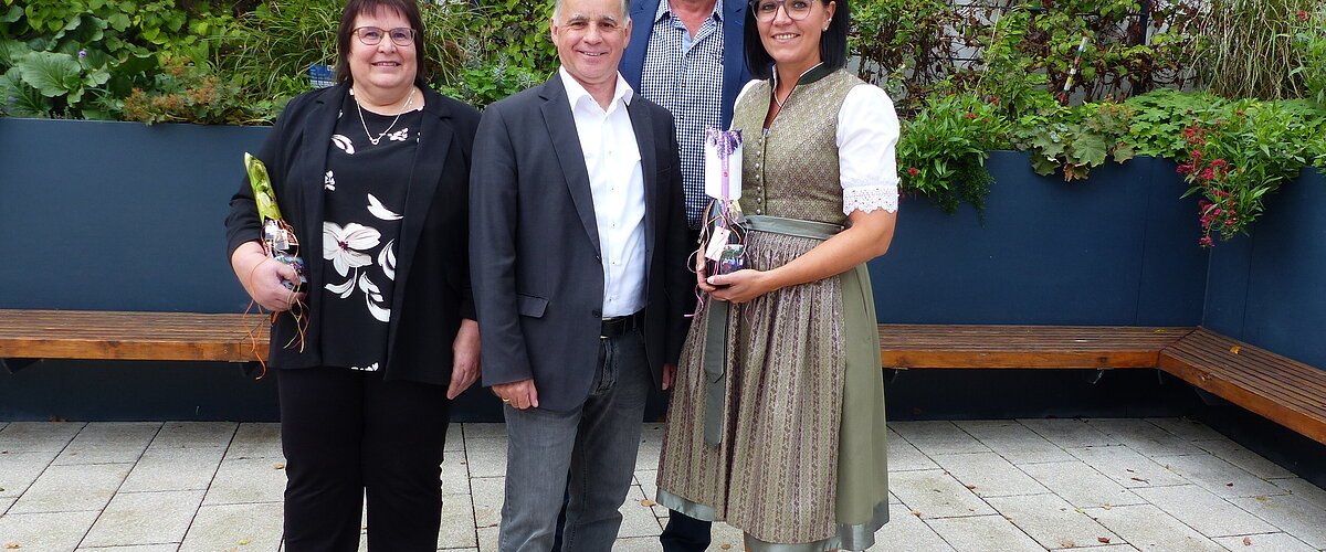 Vier Personen stehen auf gepflastertem Hof vor einem schwarzen Hochbeet mit grünen Pflanzen und Bänken entlang des Beetes, zwei Personen halten Geschenke.