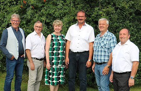 Sechs Personen stehen nebeneinander vor  grünen Bäumen, alle blicken in die Kamera und lächeln.