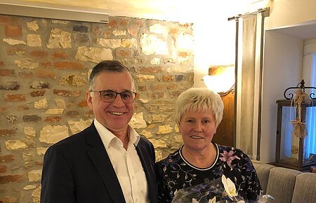 Zwei Personen, ein Mann und eine Frau stehen in einem Raum mit Steinwand im Hintergrund und einem Sitzsofe. Vor der Frau steht eine verpackte weiße Orchideenpflanze auf einem Holztisch.