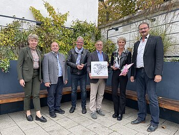 Sechs Personen stehen nebeneinander vor einer hohen Beet in anthrazit mit Pflanzen, darunter Bänke entlang des Beetes. Zwei Personen halten Geschenke in den Händen.