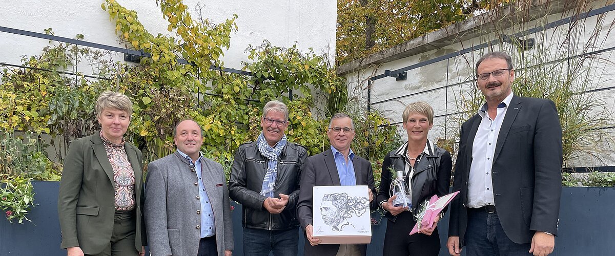 Sechs Personen stehen nebeneinander vor einer hohen Beet in anthrazit mit Pflanzen, darunter Bänke entlang des Beetes. Zwei Personen halten Geschenke in den Händen.