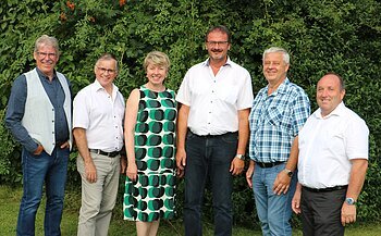 Sechs Personen stehen nebeneinander vor  grünen Bäumen, alle blicken in die Kamera und lächeln.