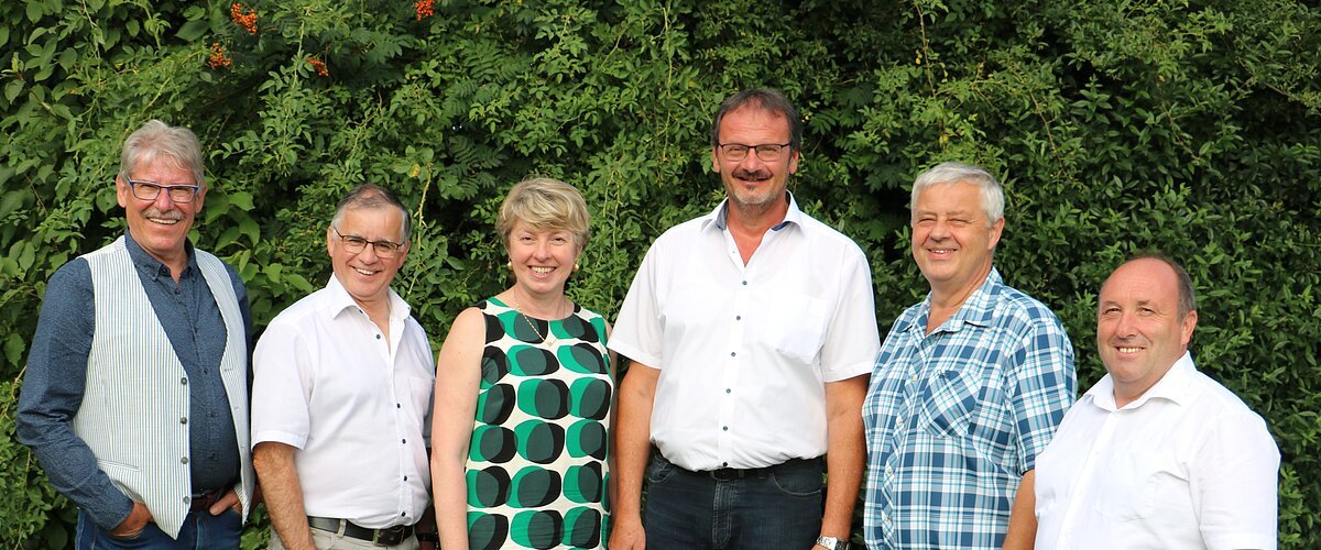 Sechs Personen stehen nebeneinander vor  grünen Bäumen, alle blicken in die Kamera und lächeln.