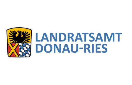Wappen des Landratsamts DONAU-RIES: auf dem Wappen oben ein schwarzer Adler auf gelbem Hintergrund. darunter links ein gelbes Kreuz auf rotem Hintergrund und rechts davon bayerisches Rautenmuster in weiß-blau. Rechts neben dem Wappen der Text: Landratsamt Donau-Ries.