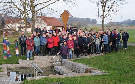 Große Gruppe älterer Menschen steht im Park vor einem kleinen Wasserbecken mit Treppe und Geländer. Monatliche Wanderung für Senioren vom Verein Soziales Miteinander
