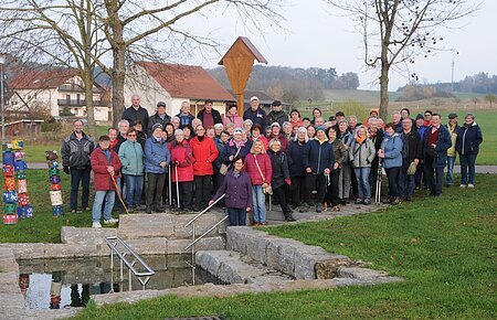 Große Gruppe älterer Menschen steht im Park vor einem kleinen Wasserbecken mit Treppe und Geländer. Monatliche Wanderung für Senioren vom Verein Soziales Miteinander