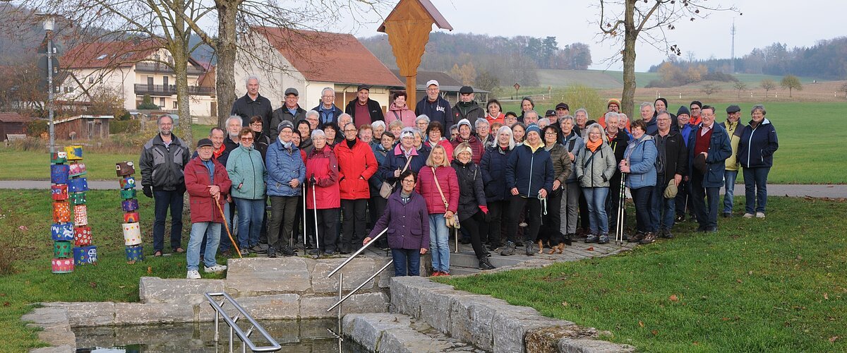 Große Gruppe älterer Menschen steht im Park vor einem kleinen Wasserbecken mit Treppe und Geländer. Monatliche Wanderung für Senioren vom Verein Soziales Miteinander