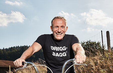 Mann mit schwarzem T-Shirt mit der weißen Aufschrift "DREGG-SAGG" und Jeans sitzt lachend auf orangefarbenem Fahrrad auf Feldweg bei Sonnenschein