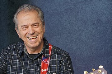 Ein älterer Mann spielt lächelnd Akustikgitarre vor einem blauen Hintergrund.