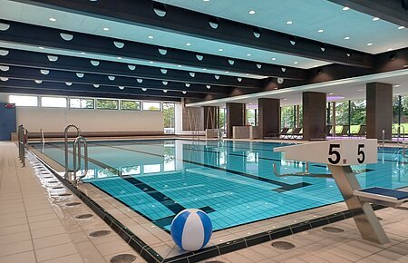 Innenaufnahme des leeren Schwimmbads - Jurabad Monheim. Zu sehen ist ein Schwimmbecken gefüllt mit strahlendem Wasser, ein Sprungbrett und ein weiß-blauer Wasserball liegt dekorativ im Vordergrund. Im Hintergrund des Hallenbads sind große Fenster zu sehen.