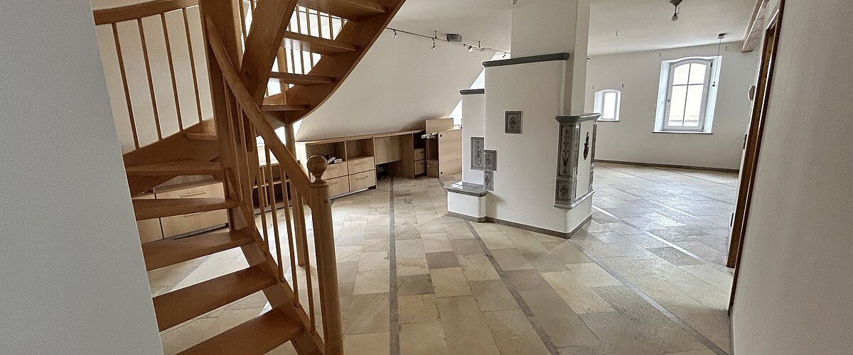Mietobjekt Maisonetten Wohnung Marktplatz 11, Monheim.
Offener Raum mit Holztreppe, Fliesenboden, weißen Wänden und zwei Fenstern in einer Dachgeschosswohnung