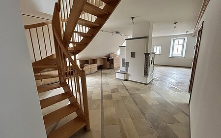 Mietobjekt Maisonetten Wohnung Marktplatz 11, Monheim.
Offener Raum mit Holztreppe, Fliesenboden, weißen Wänden und zwei Fenstern in einer Dachgeschosswohnung