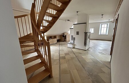 Mietobjekt Maisonetten Wohnung Marktplatz 11, Monheim.
Offener Raum mit Holztreppe, Fliesenboden, weißen Wänden und zwei Fenstern in einer Dachgeschosswohnung