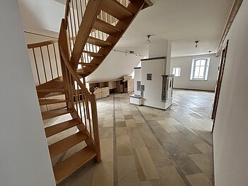 Mietobjekt Maisonetten Wohnung Marktplatz 11, Monheim.
Offener Raum mit Holztreppe, Fliesenboden, weißen Wänden und zwei Fenstern in einer Dachgeschosswohnung